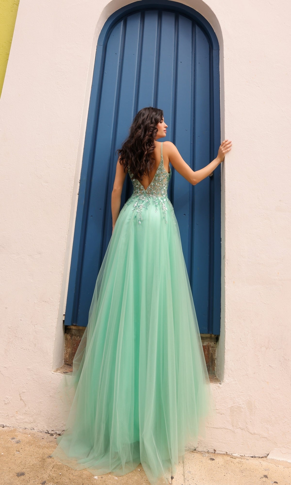 A woman in the Deep V-Back Long Tulle A-Line Prom Dress Q1391, featuring a mint green hue and embroidered flowers, stands facing a tall arched blue door, softly touching it with one hand.